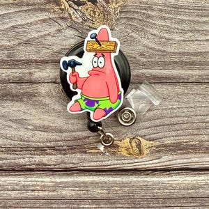 May include: A pink cartoon character with a wooden plank on its head and a hammer in its hand. The character is attached to a black retractable badge reel with a clear plastic badge holder.