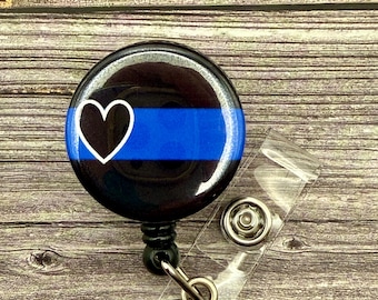 May include: A black and blue retractable badge holder with a thin blue line and a black heart. The badge holder is on a wooden surface.