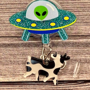 May include: A retractable badge reel featuring a teal glitter UFO with a green alien and a black and white cow charm. The UFO has yellow and blue accents. The cow charm is attached with a silver chain.