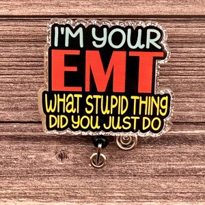 May include: A clear plastic badge holder with a glittery border. The text on the badge holder reads "I'm your EMT What stupid thing did you just do". The text is in black, yellow, and red. The badge holder has a black retractable badge reel.