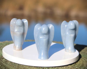 May include: Three light blue angel figurines are displayed on a white, round platform. The angels have detailed wings and are carved from a stone-like material. The background is blurred, with a glimpse of water and foliage.