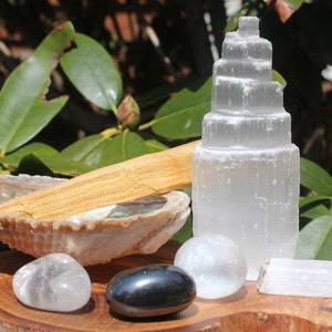 May include: A collection of crystals and tools for meditation and spiritual practice, including a selenite tower, a selenite wand, a clear quartz crystal, a black obsidian crystal, and a palo santo stick resting in a seashell.