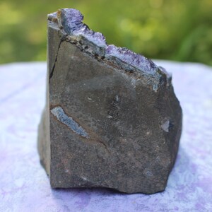 May include: A large, rough, dark gray amethyst geode with purple crystals lining the inside. The geode is sitting on a purple surface.