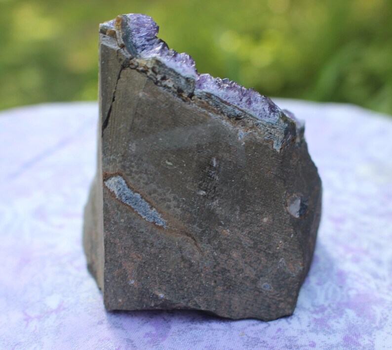 May include: A large, rough, dark gray amethyst geode with purple crystals lining the inside. The geode is sitting on a purple surface.
