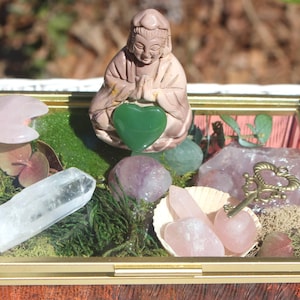 May include: A glass box filled with crystals, moss, and a small figurine of a meditating person holding a green heart. The box has a gold trim and a pink and white patterned background.