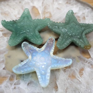 May include: Three carved starfish figurines. Two are a translucent green, and one is opalescent white and blue. The starfish are detailed with textured surfaces and are arranged on a light-colored, patterned surface.