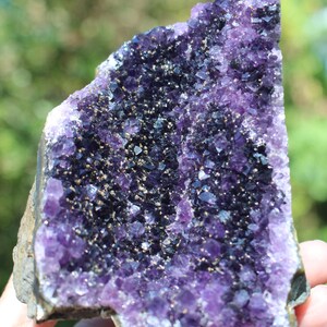 May include: A large amethyst geode with a purple and dark purple crystal cluster. The geode is partially open, revealing the interior crystals. The geode is on a rough, natural surface.
