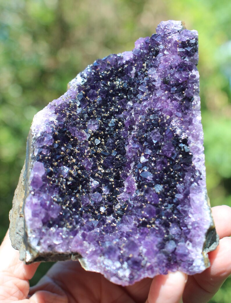 May include: A large amethyst geode with a purple and dark purple crystal cluster. The geode is partially open, revealing the interior crystals. The geode is on a rough, natural surface.