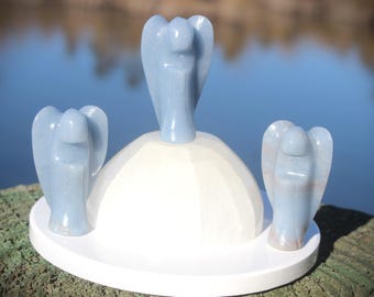 May include: A set of three light blue angel figurines on a white wooden base. The central angel is taller, with two smaller angels flanking it. The base is round with a white rim, set against a blurred water background.
