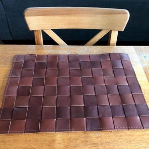 May include: A rectangular, woven leather placemat in shades of brown. The placemat is made of small, square pieces of leather woven together. The edges are stitched with white thread. The placemat is on a wooden table.