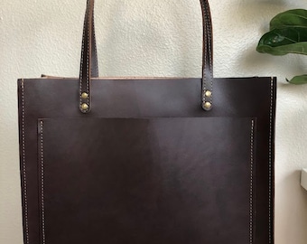 May include: A dark brown leather tote bag with a large front pocket and two top handles. The bag has a stitched design around the edges and brass rivets on the handles.