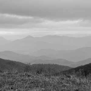 May include: A black and white photograph of a mountain range with a cloudy sky. The mountains are layered and appear to be in the distance. The sky is overcast and the light is muted.