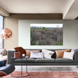 May include: A gray sofa with brown, beige, and white throw pillows in a living room with a large window, a copper pendant light, and a framed landscape photograph of a rocky cliff and trees.