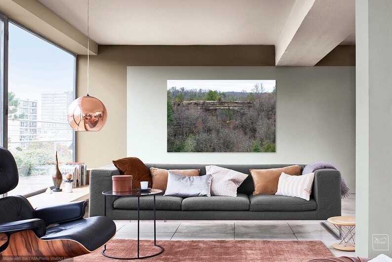 May include: A gray sofa with brown, beige, and white throw pillows in a living room with a large window, a copper pendant light, and a framed landscape photograph of a rocky cliff and trees.