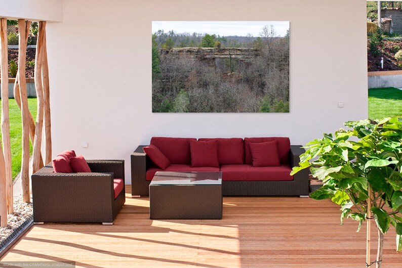 May include: A red sectional sofa with brown wicker frames and cushions sits on a wooden deck. A small brown wicker coffee table sits in front of the sofa. A green plant is in a pot on the right side of the image. A large framed photograph of a rocky cliff covered in trees hangs on the wall behind the sofa.