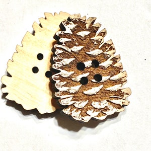 May include: Two wooden buttons shaped like pine cones. The top button has a detailed pine cone design and the bottom button is plain. Both buttons have four holes.