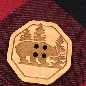 May include: A wooden button with a carved design of a bear and trees. The button has four holes for sewing.