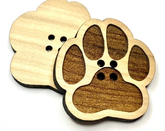 May include: Two wooden buttons, one shaped like a flower and the other shaped like a paw print. Both buttons have a brown wood grain and are unfinished.
