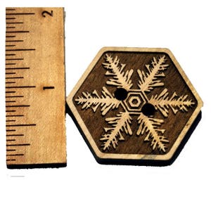 May include: A wooden button with a snowflake design. The button is hexagonal and has two holes for sewing. The snowflake is carved into the wood and is a light brown color.