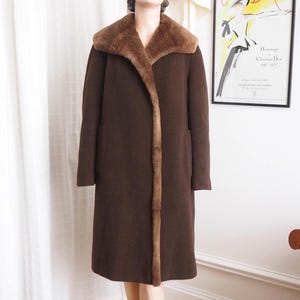 Puede incluir: Un abrigo de lana marrón con cuello y ribete de piel sintética, expuesto en un maniquí. El abrigo llega a la rodilla y se combina con un sombrero a juego. El maniquí lleva zapatos negros. Un póster de moda vintage es visible en el fondo.