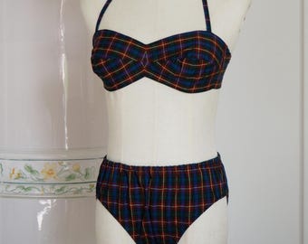 May include: A two-piece swimsuit featuring a dark blue, green, and red plaid pattern with a halter-style top and high-waisted bottoms. A straw hat with embroidered yellow, blue, and purple floral details rests on the mannequin head above the set.