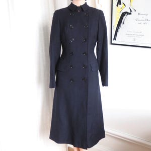 Puede incluir: Un abrigo azul marino de doble botonadura con un diseño clásico. El abrigo presenta un corte entallado, un cuello y un dobladillo hasta la rodilla. Tiene dos filas de botones oscuros y dos bolsillos delanteros. El abrigo se exhibe en un maniquí con tacones negros.