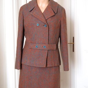 Peut inclure: Un tailleur deux pièces vintage en tweed marron et bleu moucheté. La veste présente un design croisé avec trois boutons bleus et une jupe assortie. Le costume est présenté sur un mannequin.