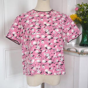 May include: A pink short-sleeved top with black trim, patterned with white and black objects, including hats and plates. The top is displayed on a mannequin. The top is a vintage style.