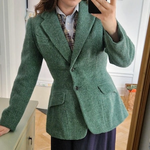 May include: A textured green blazer with a notched lapel, two front buttons, and two flap pockets. The blazer is worn over a light blue collared shirt, a brown and white plaid waistcoat, and a solid dark navy blue skirt that reaches below the knees.