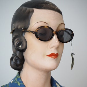 Puede incluir: Un maniquí de estilo vintage con el pelo oscuro peinado en ondas, lleva una camisa azul con estampado floral y gafas de sol ovaladas de carey con un cordón negro.