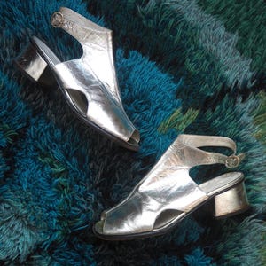 May include: A pair of silver open-toe sandals with a block heel. The sandals have a strap around the ankle and a buckle closure. The shoes are displayed on a textured, multi-colored rug.