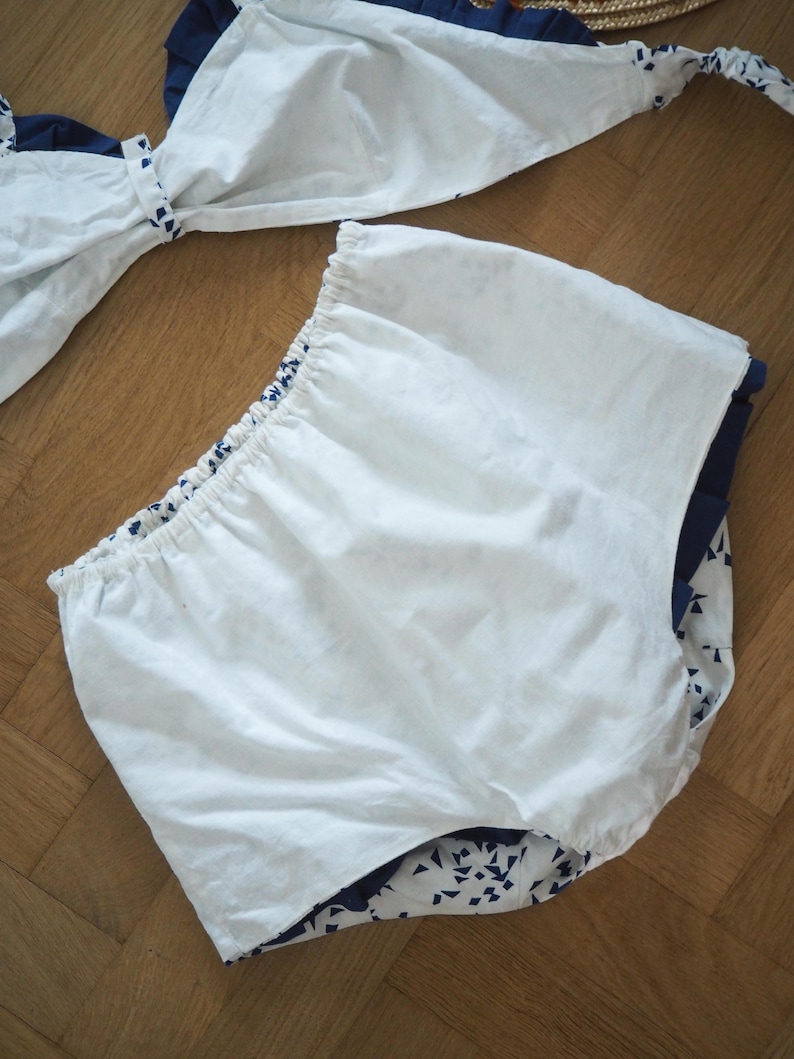 May include: White and navy blue two-piece swimsuit set. The top features a bow detail, and the bottoms are high-waisted. The navy blue fabric has a white geometric pattern. The swimsuit is made of a lightweight fabric.