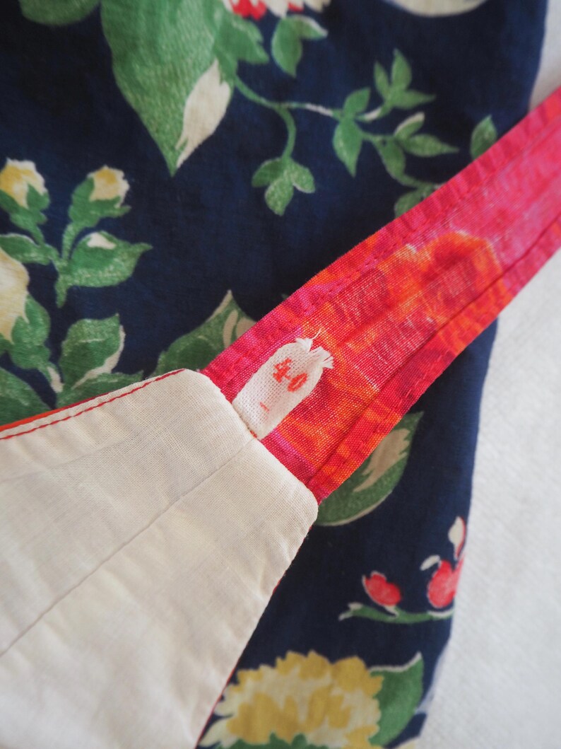 May include: Close-up of fabric with a floral pattern in navy blue, green, and yellow. A bright pink strap with orange accents is visible, along with a white fabric edge. The strap has a small, faded text.
