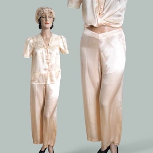 May include: A vintage peach-colored satin pajama set with lace trim. The top has short sleeves and a button-up front. The pants have a wide leg.