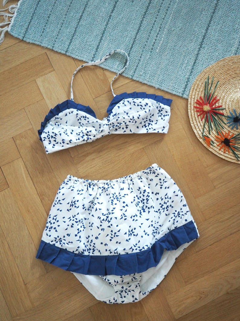 May include: A white and blue patterned bikini set with a matching high-waisted bottom. The bikini top has a ruffled edge and halter straps. The bottom also features a ruffled edge. A straw hat with colorful embroidered flowers is also present.