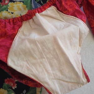 May include: Close-up of a red and pink floral patterned garment with a white interior lining. The garment has a gathered elastic waistband and red stitching. A dark blue floral fabric is visible in the background.