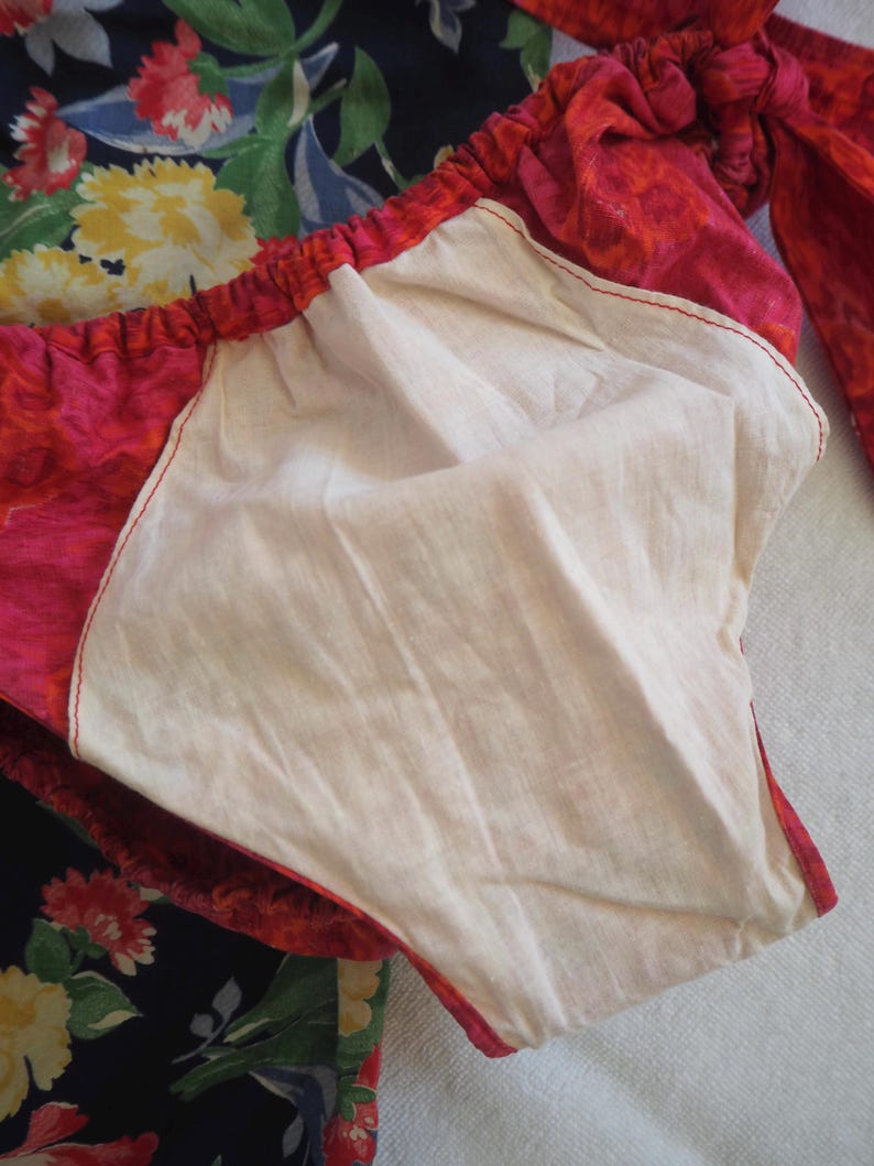 May include: Close-up of a red and pink floral patterned garment with a white interior lining. The garment has a gathered elastic waistband and red stitching. A dark blue floral fabric is visible in the background.