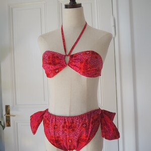 May include: A two-piece bikini set in shades of red and pink, displayed on a mannequin. The halter top features a central ring detail, and the bottoms have side ties. The fabric has a floral pattern.