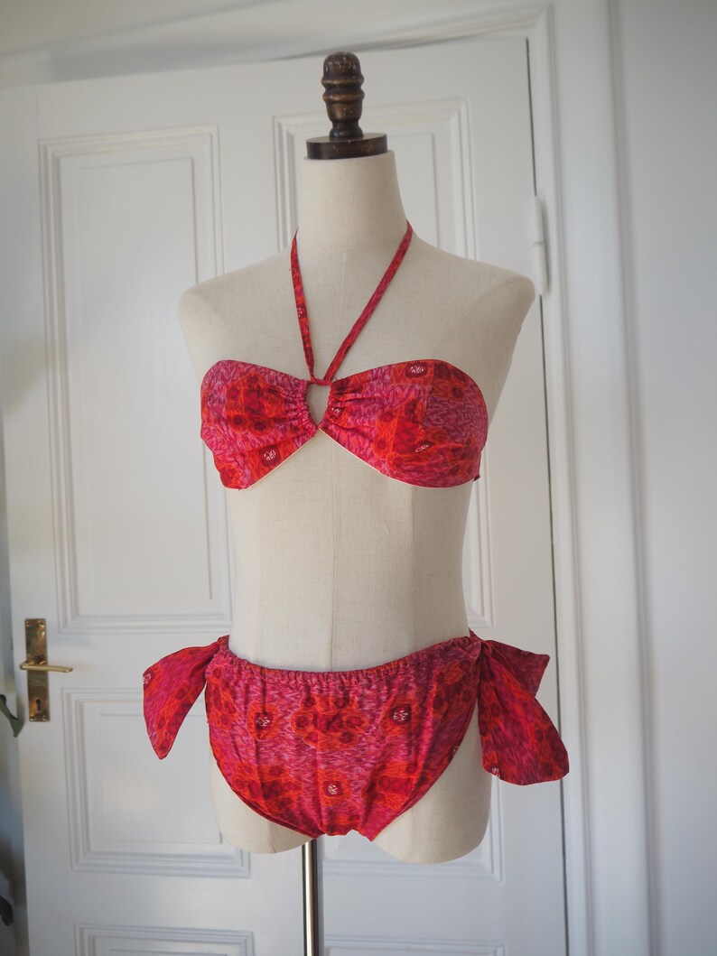 May include: A two-piece bikini set in shades of red and pink, displayed on a mannequin. The halter top features a central ring detail, and the bottoms have side ties. The fabric has a floral pattern.