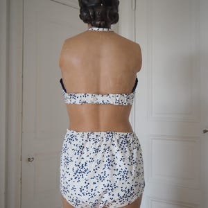 May include: A two-piece swimsuit displayed on a mannequin. The top is a halter style with a white base and a blue floral pattern. The bottoms are high-waisted with the same blue floral design. The mannequin has dark hair and is positioned in front of a white door.