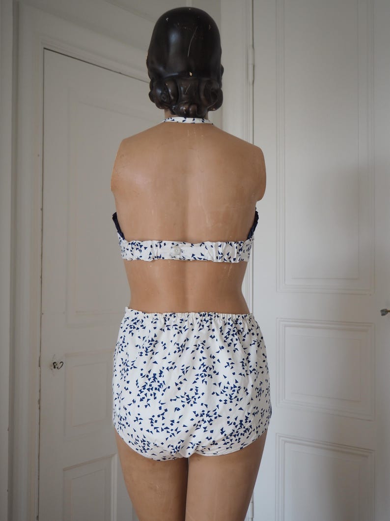 May include: A two-piece swimsuit displayed on a mannequin. The top is a halter style with a white base and a blue floral pattern. The bottoms are high-waisted with the same blue floral design. The mannequin has dark hair and is positioned in front of a white door.