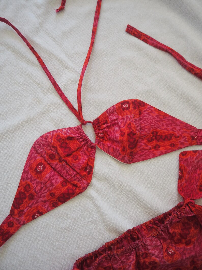 May include: A red and pink patterned bikini set. The top is a halter style with adjustable ties, and the bottoms are a low-rise design. The fabric features a repeating floral pattern in shades of red and pink.