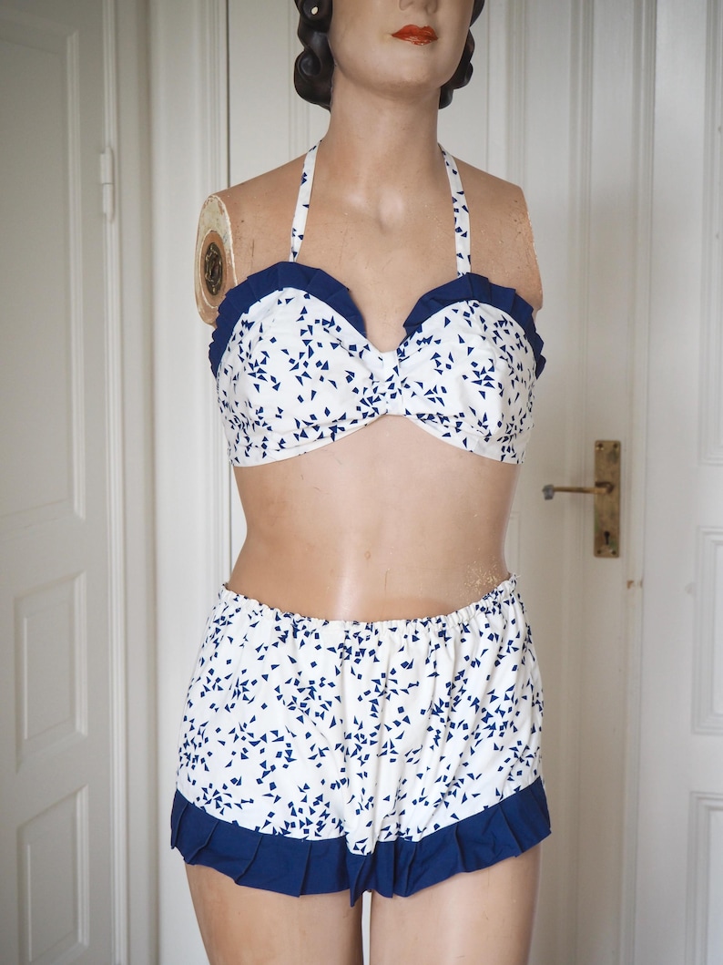 May include: A vintage-style two-piece swimsuit. The halter-neck top and high-waisted shorts are white with a blue geometric pattern. The edges of the top and shorts are trimmed with a solid navy blue ruffle. The swimsuit is displayed on a mannequin.