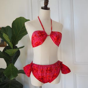 May include: A vintage two-piece swimsuit in shades of red and pink, displayed on a mannequin. The halter-style top and high-waisted bottoms feature a floral pattern. The bottoms have side ties.