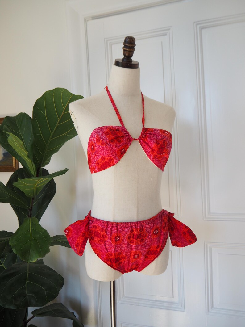 May include: A vintage two-piece swimsuit in shades of red and pink, displayed on a mannequin. The halter-style top and high-waisted bottoms feature a floral pattern. The bottoms have side ties.
