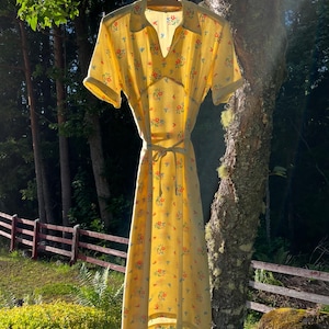 May include: A yellow floral print dress with short sleeves and a tie waist hangs from a wooden hanger. The dress features a V-neck collar and is adorned with small red, blue, and yellow flowers. The dress is hanging outside in front of a forest.