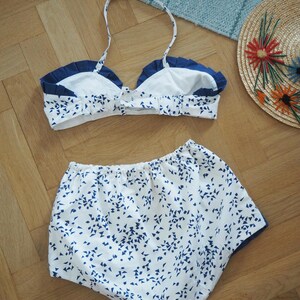 May include: A white and blue patterned bikini set with a halter top and high-waisted bottoms. The bikini top has a navy blue trim and the bottoms have a navy blue trim. A straw hat with colorful embroidered flowers is also in the image.