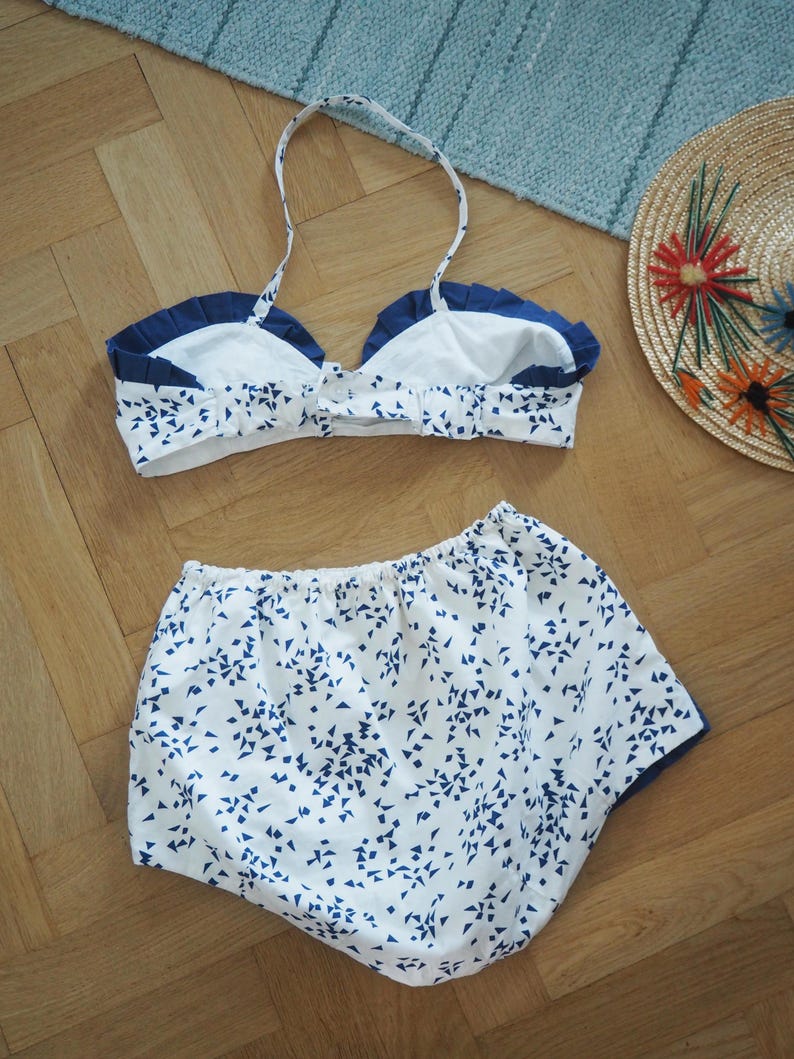 May include: A white and blue patterned bikini set with a halter top and high-waisted bottoms. The bikini top has a navy blue trim and the bottoms have a navy blue trim. A straw hat with colorful embroidered flowers is also in the image.