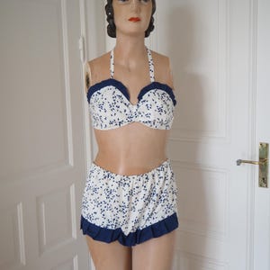 May include: A vintage-style two-piece swimsuit. The halter-neck bikini top and high-waisted shorts are white with a blue floral pattern. The edges of the top and shorts are trimmed with a dark blue ruffle.