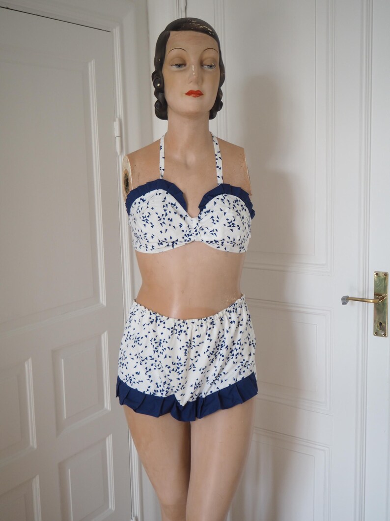 May include: A vintage-style two-piece swimsuit. The halter-neck bikini top and high-waisted shorts are white with a blue floral pattern. The edges of the top and shorts are trimmed with a dark blue ruffle.